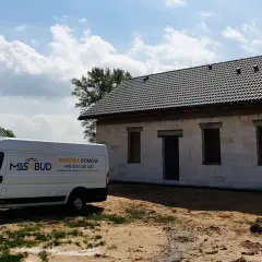 Biały van budowlany M&S BUD przed niedokończonym domem z szarej cegły pod czarnym dachem.
