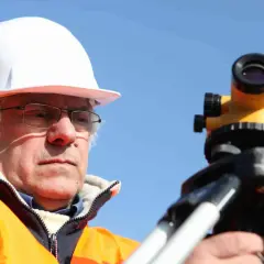Geodeta w kasku i kamizelce odblaskowej pracuje z niwelatorem na budowie.