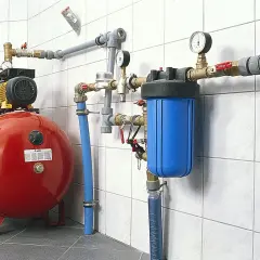Czerwony zbiornik przeponowy, pompa, niebieski filtr wody i instalacja rurowa na białej kafelkowej ścianie.