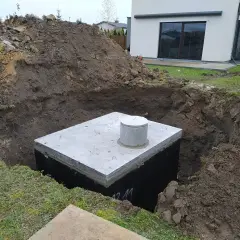 Betonowy zbiornik na deszczówkę w wykopie, otoczony ziemią.