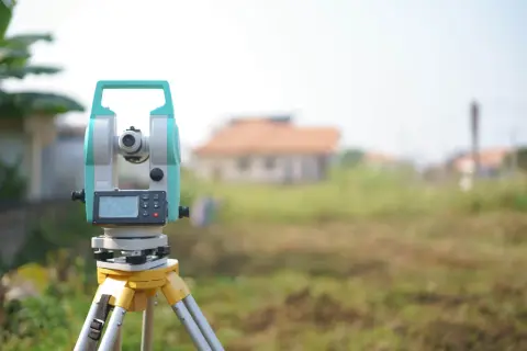 Geodezyjny tachimet na statywie na tle niezabudowanej działki i domu.