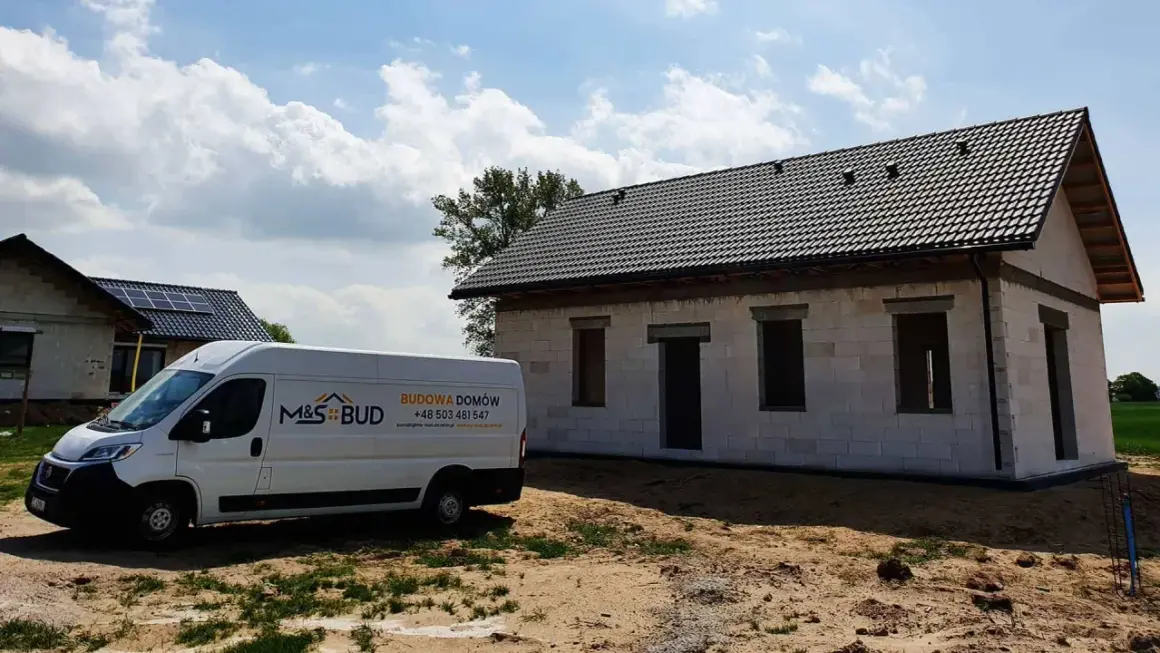 Biały van budowlany M&S BUD przed niedokończonym domem z szarej cegły pod czarnym dachem.