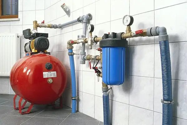 Czerwony zbiornik przeponowy, pompa, niebieski filtr wody i instalacja rurowa na białej kafelkowej ścianie.